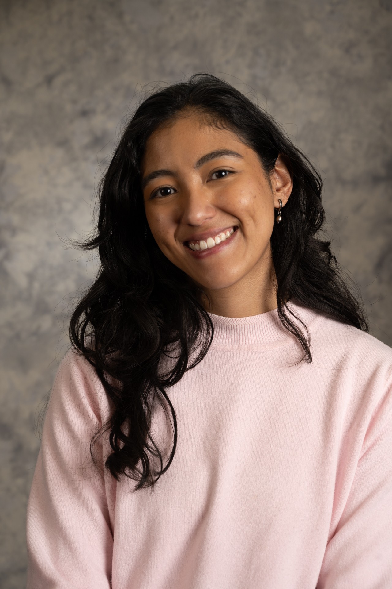 Image of woman smiling. She has dark brown hair and is wearing a pink sweater and earrings. 