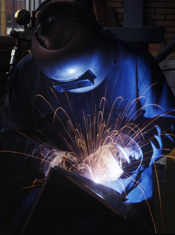 A person wears a welding helmet, suit, and gloves while welding. Sparks are flying as they work