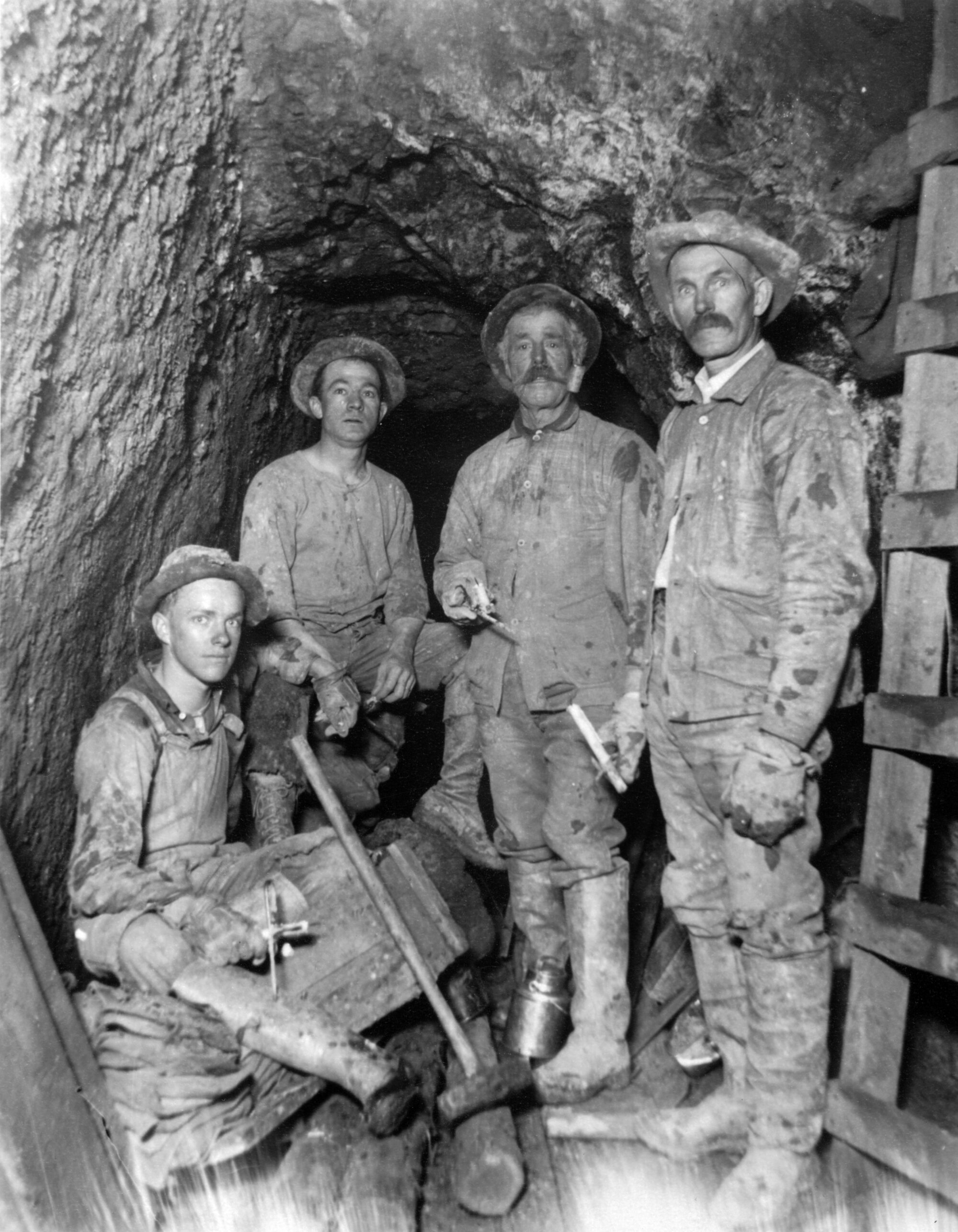 Four men stand in a mining tunnel, looking at the camera. They wear work clothes and hold mining tools such as a hand-drill and sledge hammer.