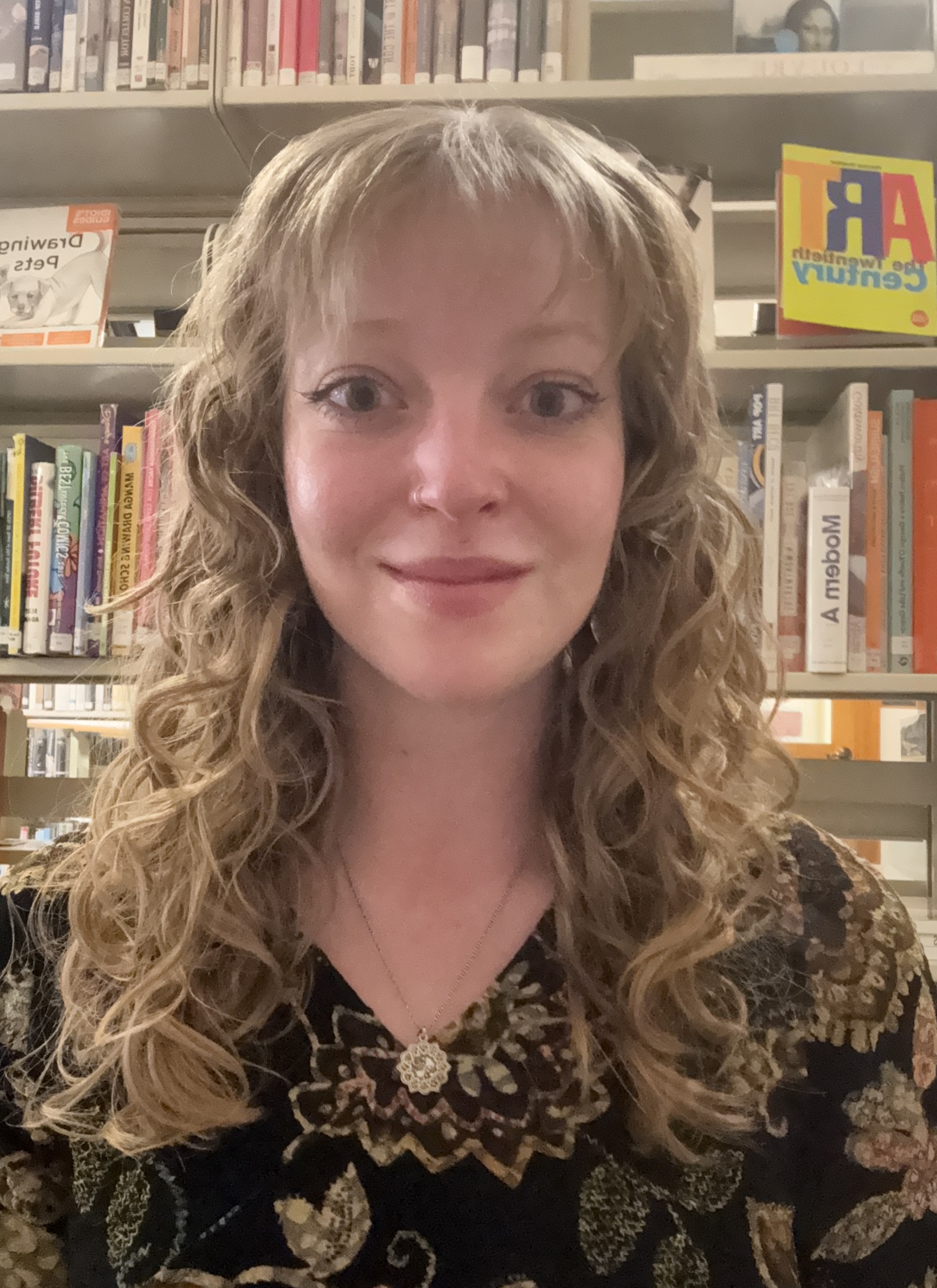 Rebecca Goddard, a white wman with curly blonde hair and blue eyes, smiles at the camera while standing in front of a library book shelf.