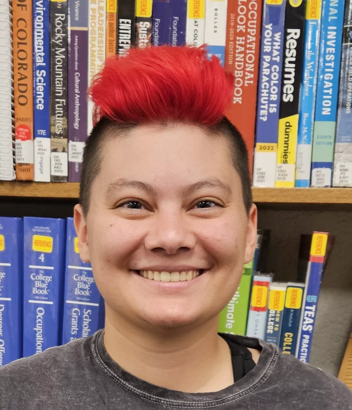Image of person in front of library books. They have a short, red mohawk, and they are smiling.