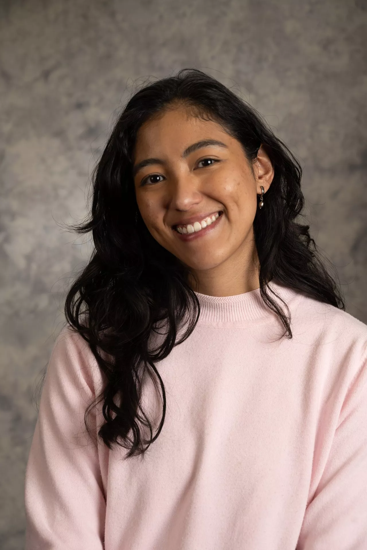 Image of woman smiling. She has dark brown hair and is wearing a pink sweater and earrings. 
